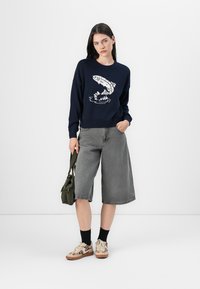 Mornarsko pleteno pulover s grafiko rib, sivi denimski culotte, črne nogavice in bež superge z živalskim vzorcem. Olivno zelena torba čez ramo.