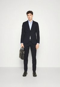 Tmavomodrý oblek, upravený so sakom a nohavicami úzkeho strihu. Doplněný svetlomodrou košeľou a čiernymi koženými topánkami, držiaci čiernu tašku.