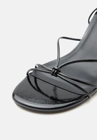 Μαύρο σανδάλι από δερματίνη με λεπτές, γυαλιστερές ράβδους δεμένες σε έναν μικρό κόμπο πάνω από το ανοιχτό δάχτυλο σε ουδέτερο φόντο.