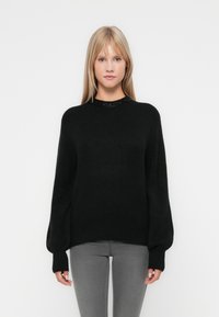 Pull en tricot noir avec un col rond, des manches larges et des poignets côtelés. Porté sur un jean gris, avec une texture lisse et une coupe décontractée.