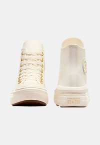 Zapatillas altas de lienzo blanco con ojales dorados, suela de goma y parche de logo en contraste en el lateral. Textura suave y punta redondeada.