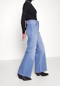 Persona che indossa jeans blu a vita alta e gamba larga, top a maniche lunghe nero, stivali neri con tacco e una collana di perline, in posizione di profilo.