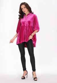 Blusa di velluto fucsia con chiusura a bottoni, vestibilità comoda e orlo arrotondato, abbinata a pantaloni neri aderenti e tacchi aperti.