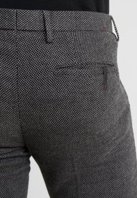 Šedé kalhoty s texturou a jemným puntíkovaným vzorem, předními i zadními kapsami a volitelnými zipy s minimalistickými kovovými detaily.