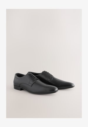Chaussures habillées en cuir noir à texture lisse, bout arrondi et design à lacets. Dotées d'un talon bas et d'accents de couture minimalistes.