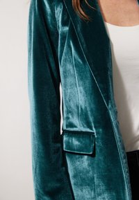 Teal sametový blazer s hladkou texturou, přiléhavým střihem, klopami a jednou náprsní kapsou, nošený přes bílý top.