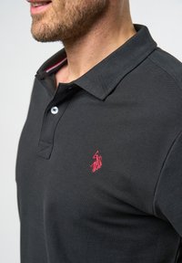 Svart pikétröja med strukturerad tyg, med krage, två-knapps stängning och en röd broderad logotyp på vänster bröst.