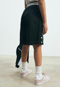 En person iført sorte knælange shorts og hvide sneakers holder en sort og hvid jakke mod en ensfarvet baggrund.