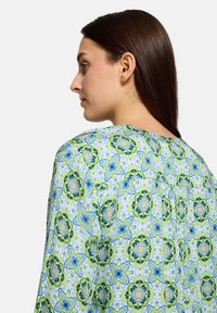 Bluzka w wzór z zielonym i niebieskim motywem kwiatowym, miękka tkanina, okrągły dekolt i luźny krój. Widoczne szwy i detal z tylnym karczkiem.