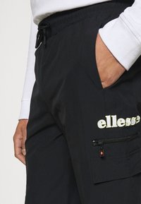 Svarta byxor med elastisk midja och sidoficka, med en gul "ellesse"-logga och en ficka med dragkedja på låret. Slät tygtextur.