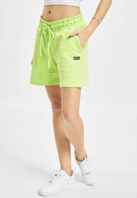Neon-grüne Baumwollshorts mit einem elastischen Bund, verstellbarem Kordelzug, Seitentaschen und "DEF" in Schwarz auf dem linken Bein gedruckt.