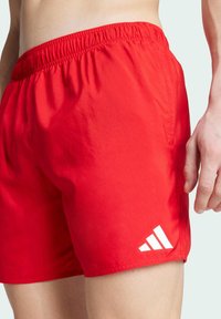 Červené plavecké šortky s elastickým pásom. Na spodnej strane sa nachádza biele logo Adidas. Materiál vyzerá hladko a je ľahký.
