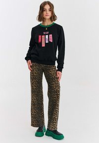 Černá mikina s barevnými textovými nášivkami, kombinovaná s volnými kalhotami s leopardím vzorem a zelenými platformovými botami. Ležérní městský outfit.