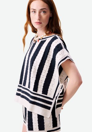 Femme aux longs cheveux roux portant un haut en maille rayé noir et blanc et un short assorti, accessoirisés d'un collier de perles.