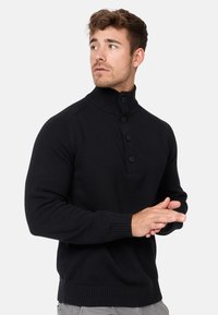 Sort striktrøje med høj krave, tre-knaps lukning, ribbede manchetter og kant. Stoffet ser blødt og tekstureret ud. Modelens hænder er foldet.