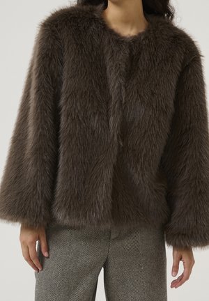Personne portant une veste en fausse fourrure marron foncé avec des manches larges et un pantalon gris à motif chevrons, debout devant un fond uni.