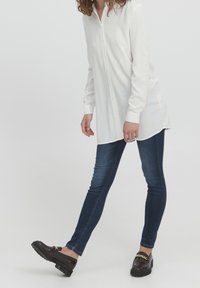 Weißes Hemd mit langen Ärmeln, kombiniert mit dunkelblauen Skinny Jeans und schwarzen Slippern mit goldener Kettenverzierung.
