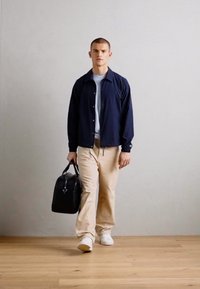 Man walking indoors wearing a navy jacket, beige pants, white sneakers, carrying a black leather duffel bag in his right hand.