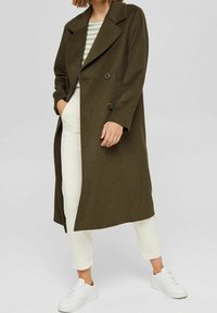 Osoba ubrana w długi, ciemnozielony, dwurzędowy wełniany płaszcz na jasną w paski koszul�ę, białe spodnie i białe trampki, stojąca z rękami w kieszeniach.