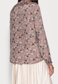 Blouse en dentelle à manches longues de couleur marron clair avec un motif de feuilles noir et blanc. Présente un col rond et des poignets froncés.