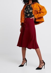 Nainen, jolla on päällään sinapinkeltainen takki, musta kukkakuosiset paita, burgundin värinen napitettu midi-hame ja mustat nauhakengät, seisoo vaaleataustalla.