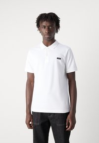 Weißes Poloshirt aus Baumwolle mit klassischem Kragen und einer Drei-Knopf-Leiste. Verfügt über ein kleines schwarzes Logopatch auf der linken Brust.