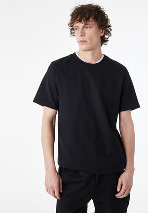 Joven con pelo rizado que lleva una camiseta negra lisa y pantalones negros, mirando hacia su izquierda sobre un fondo blanco.