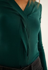 Top verde a maniche lunghe con trama a costine, caratterizzato da un design intrecciato al collo e una silhouette aderente. Abbinato a una fascia in vita scura.