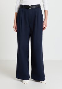 Weit geschnittene marineblaue Hose aus glattem Stoff, mit vorderen Falten und einem schwarzen Gürtel. Kombiniert mit weißen, spitzen Absätzen für einen eleganten Look.