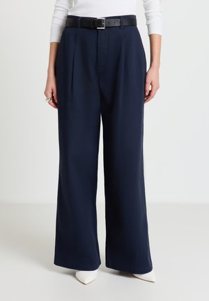 Pantalon large bleu marine en tissu lisse, avec plis sur le devant et une ceinture noire. Associé à des talons blancs pointus pour un look épuré.