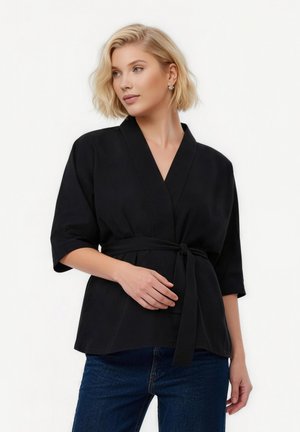 Mujer rubia con blusa estilo kimono negra con cinturón, mangas hasta el codo y vaqueros azules, mirando ligeramente hacia la izquierda.