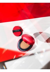 Rode cushion compact met glanzend deksel, voorzien van een zwarte rand en een ronde applicator. Open, met zicht op de sponsstructuur binnenin. Modern ontwerp.