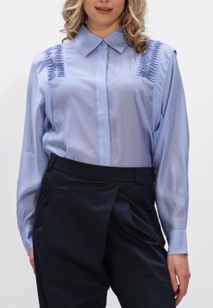 Vrouw draagt een lichtblauw blouse met lange mouwen, geplooide details op de schouders en een hoge taille zwarte broek, staand met één hand zichtbaar.