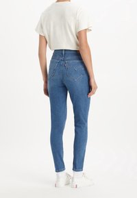Jeans de mezclilla de cintura alta en azul, con un silueta ajustada y bolsillos traseros en forma de corazón. Combinados con una camiseta corta de color beige.