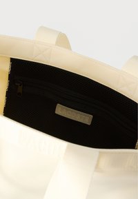 Τσάντα ώμου σε κρεμ χρώμα με κρεμ λαβές και μαύρη εσωτερική θήκη με φερμουάρ που φέρει την ετικέτα "Lacoste", με διακοσμητική ταινία με λογότυπο κατά μήκος της εσωτερικής άκρης.