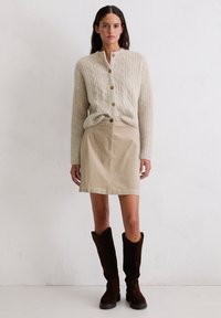 Beige Strickjacke mit Rundhalsausschnitt und braunen Knöpfen, kombiniert mit einem Khaki-Rock und braunen kniehohen Stiefeln.