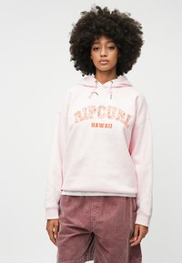 Hoodie rose avec "RIP CURL HAWAII" imprimé en orange et blanc, doté d'une capuche à cordon de serrage et d'un tissu doux. Associé à un short en velours côtelé rose.
