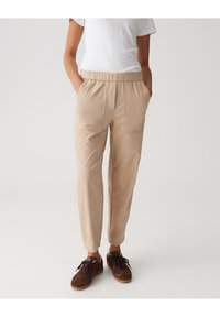 Pantalons beiges, fuselés, avec une taille élastique et des poches latérales ; fabriqués dans un tissu lisse. Associés à des baskets brunes à lacets.