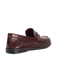 Burgundy leather loafer, featuring a textured finish, gold accent, and a flat black rubber sole. The design includes visible stitching and pleats.