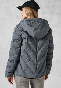 Doudoune grise avec un motif en chevron, capuche et poignets côtelés. Portée avec une casquette noire et associée à un jean gris clair.