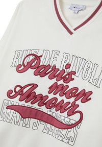 Felpa bianca con scollo a V, caratterizzata da testo ricamato color bordeaux: "Paris mon Amour" e "Rue de Rivoli Champs-Élysées." Bordo a costine in contrasto.