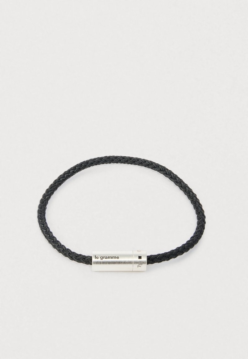 Pulseira de couro trançado preta com um fecho cilíndrico prateado gravado com "le gramme" e "7g" sobre um fundo claro.