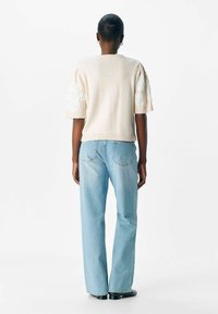 Person med kort hår i en krämfärgad stickad tröja med texturerade ärmar och ljusblå raka jeans, stående mot en enfärgad vit bakgrund.