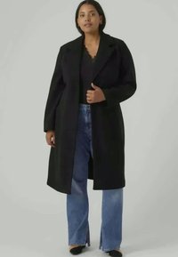 Cappotto di lana nero con rever a punta, maniche lunghe e taglio dritto, indossato sopra un top nero e jeans a gamba larga blu. Texture morbida.