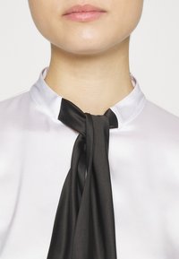 Gros plan d'une personne portant un chemisier en satin blanc à col montant et un foulard en satin noir noué de manière lâche autour du cou.