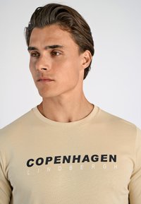 Beige långärmad skjorta med rund halsringning, prydd med djärv svart och vit text "COPENHAGEN LINDENBERGH." Slät textur, kroppsnära design.