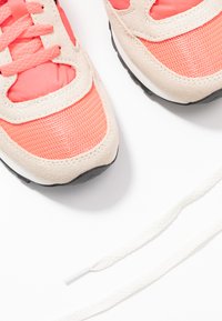 Korallenfarbene und beige Sneaker verfügen über ein Obermaterial aus Mesh mit Suede-Akzenten, einer runden Zehenpartie und einer dicken schwarzen Gummisohle. Weiße Schnürsenkel liegen neben den Schuhen.