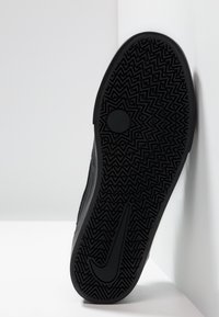 Svart sneaker med en texturerad gummisula som har zigzag-mönster och en framträdande logotyp. Sido-profil lutad mot en vit bakgrund.