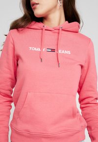 Sudadera rosa de tela suave, con un bolsillo tipo canguro, cordones y el logo "TOMMY JEANS" en blanco con un acento de estampado.