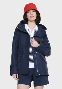 Marine vandtæt jakke med store lommer, hætte og lynlås. Bæres over en hvid og marineblå stribet skjorte med marineblå shorts. Lys rød bøttehat.